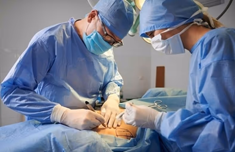 Two surgeons in blue scrubs and masks marking a patient's skin before surgery in an operating room.