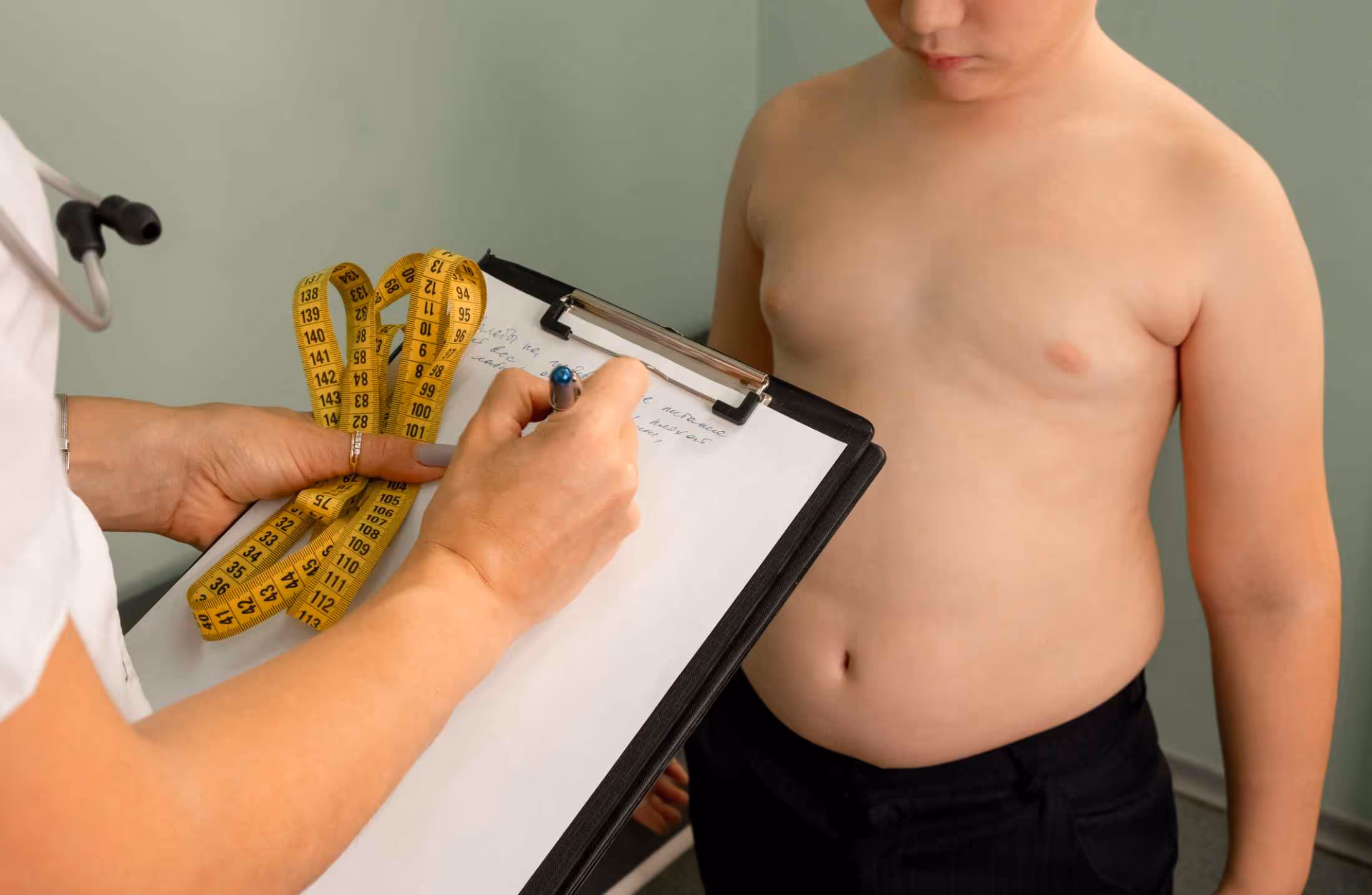 Healthcare professional holding a clipboard and measuring tape, recording notes next to a shirtless child.