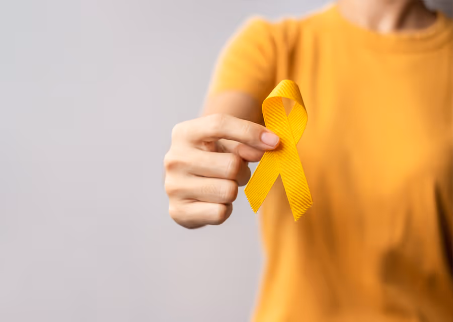 Hand holding a yellow awareness ribbon against a plain gray background, person in yellow shirt