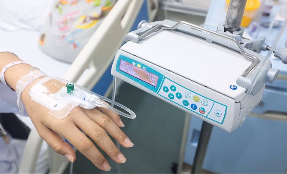 Patient's hand with IV drip attached, connected to an infusion pump in a hospital bed.
