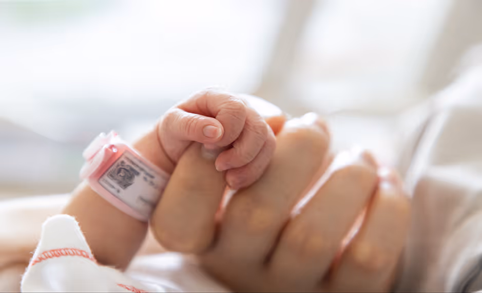 Newborn baby grasping an adult's finger, wearing a hospital ID wristband.