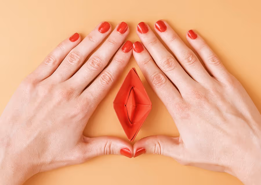 Hands with red nail polish surrounding red origami shaped like a leaf on beige background.