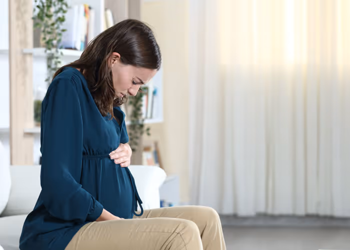 Pregnant woman sitting, holding her belly with a concerned expression in a living room.