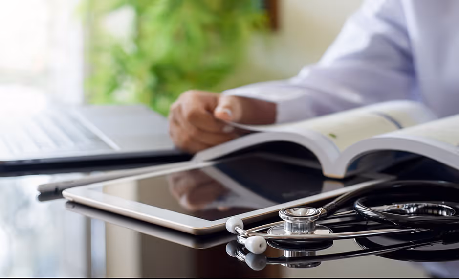 Tablet and stethoscope on table with person reading a medical book in the background.