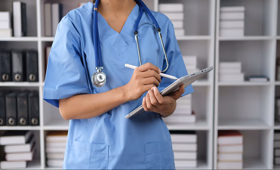 Healthcare professional in blue scrubs with stethoscope holding a tablet and stylus in a medical office.