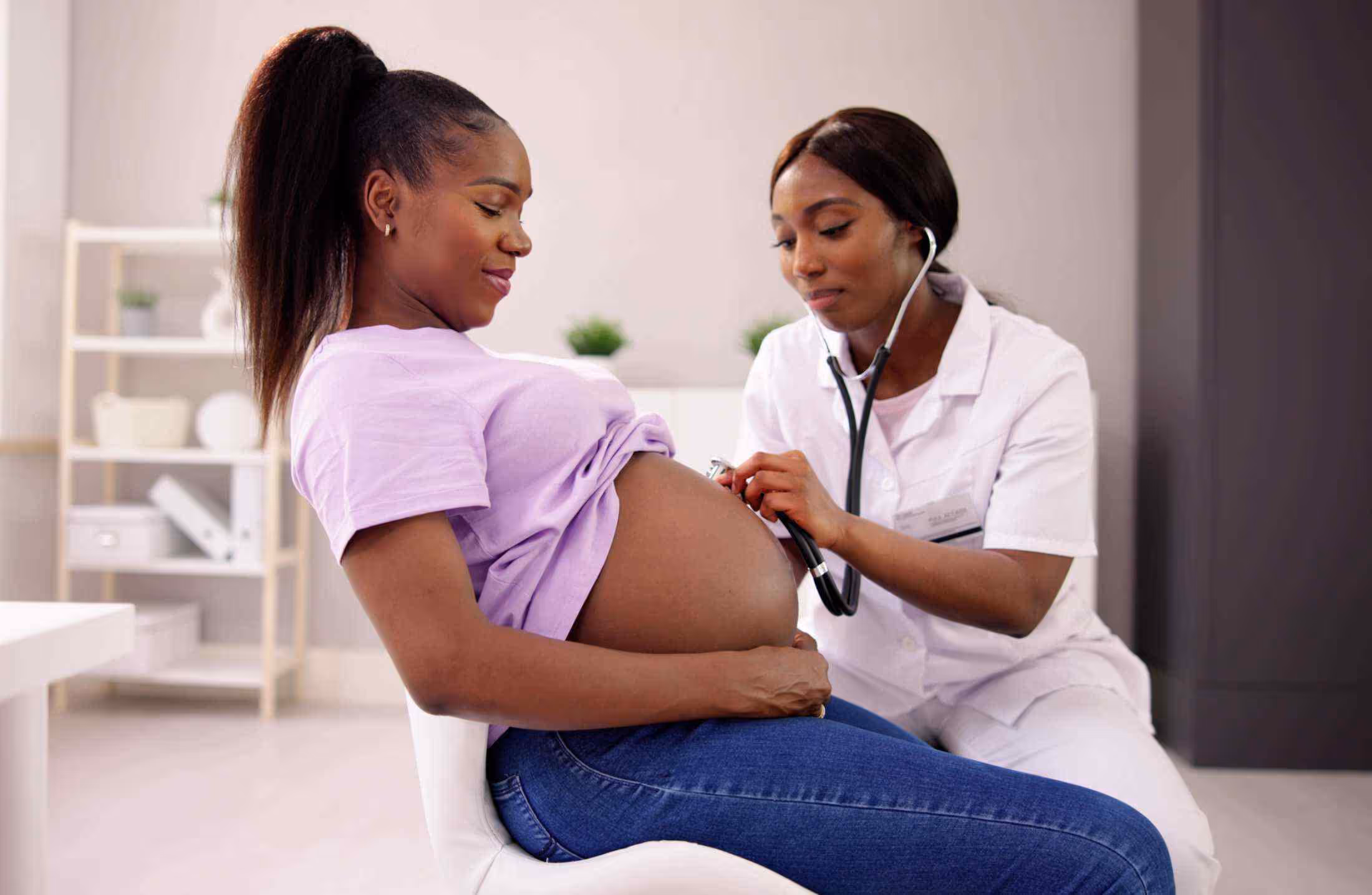 Pregnant woman in purple shirt examined by female doctor using stethoscope on her belly.