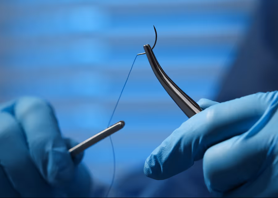 Gloved hands threading a curved surgical needle with black thread against a blue background.