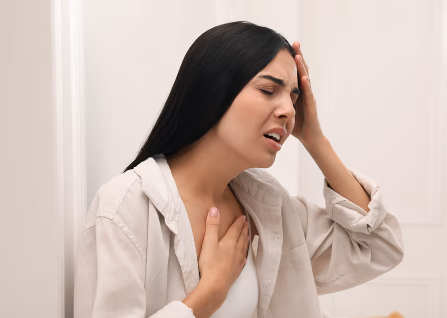Woman with long dark hair holding chest and head in pain, leaning against a white wall.