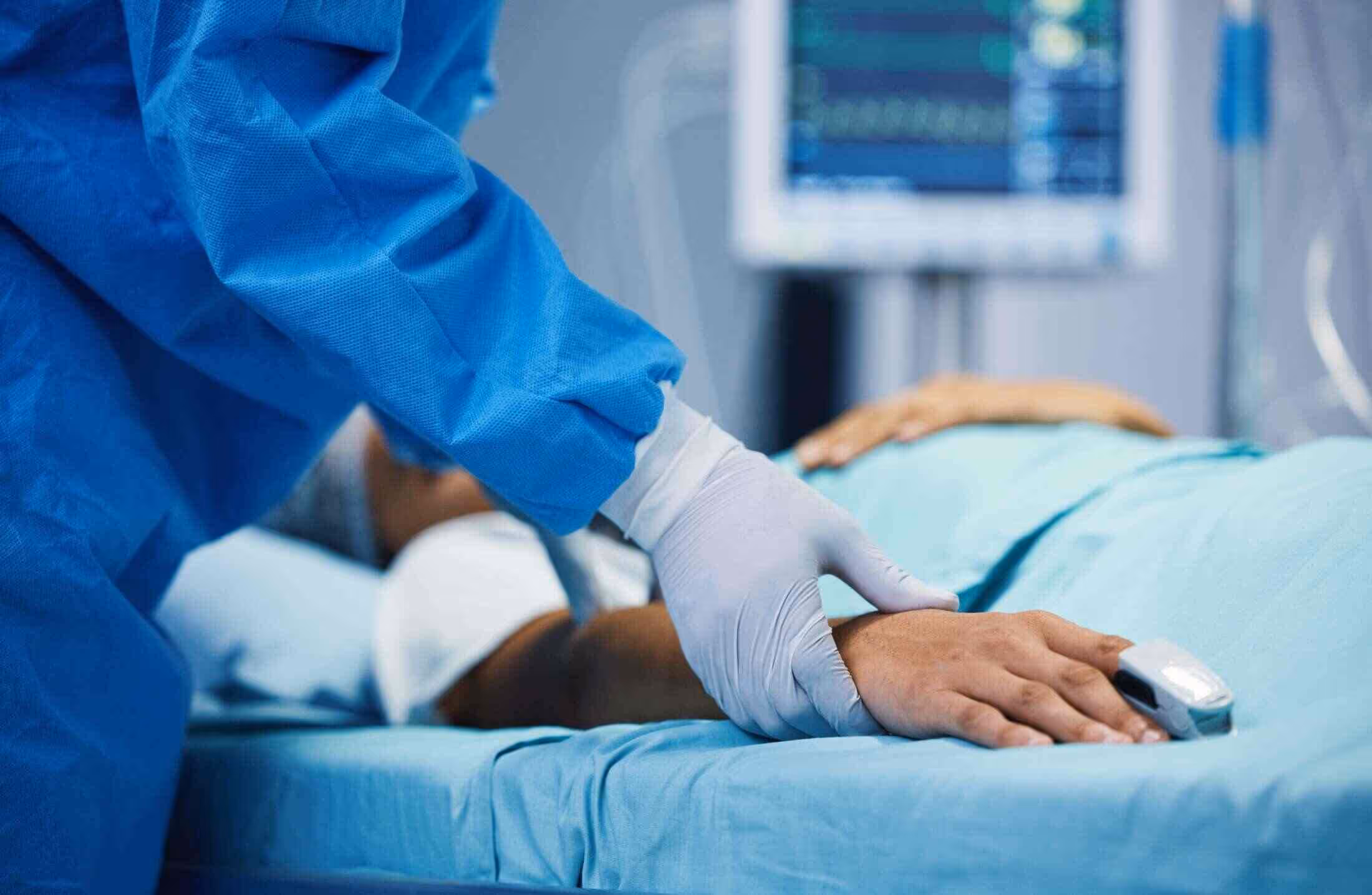 Gloved hand of healthcare worker holding patient’s hand on hospital bed with monitoring device on finger.