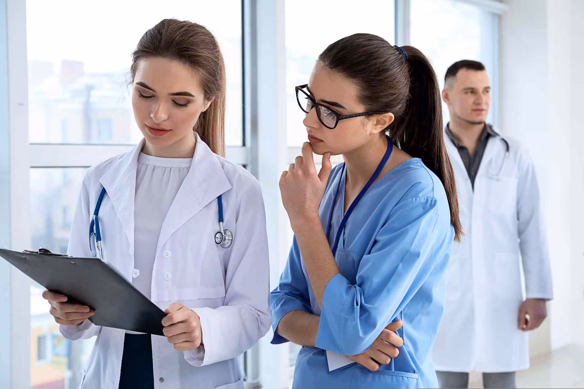 Two female medical professionals review a clipboard while a male doctor stands in the background.