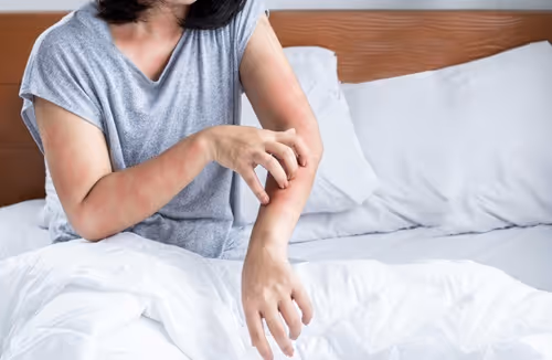 Person scratching red, irritated skin on their left forearm while sitting on a bed with white sheets.