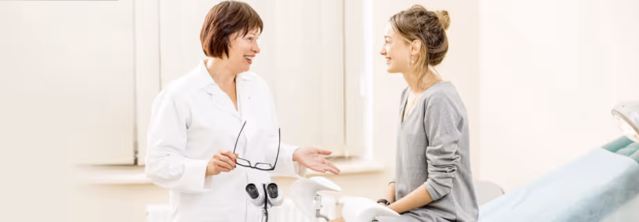 Female doctor talking and smiling with a seated female patient in a bright medical exam room.