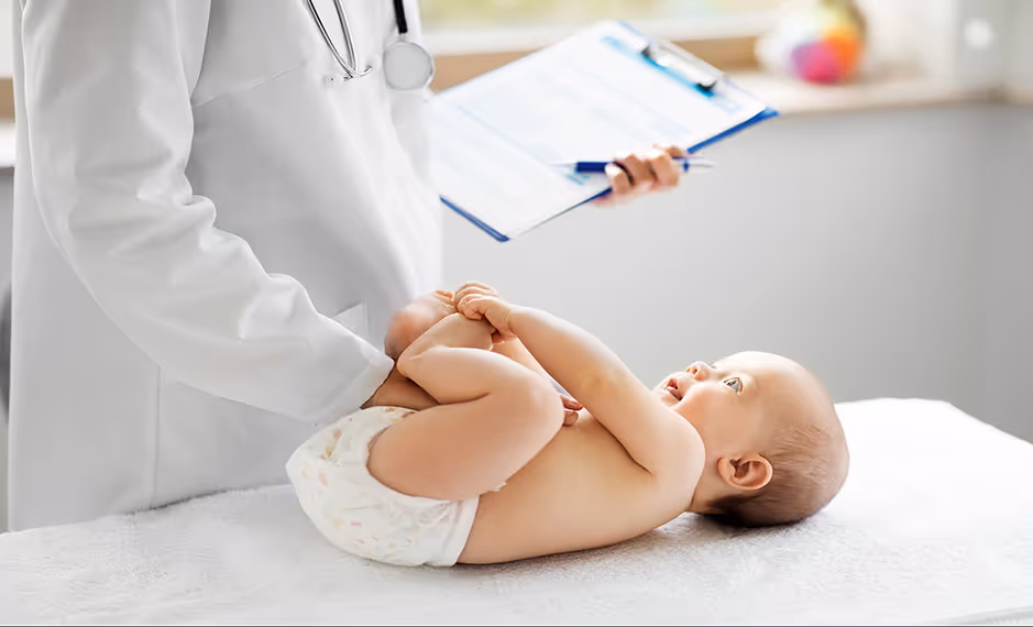 Infant lying on a table while a doctor in a white coat holds the baby's feet and writes on a clipboard.