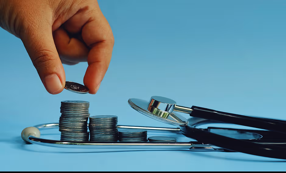 Hand stacking coins beside a stethoscope on a blue background symbolizing healthcare costs.