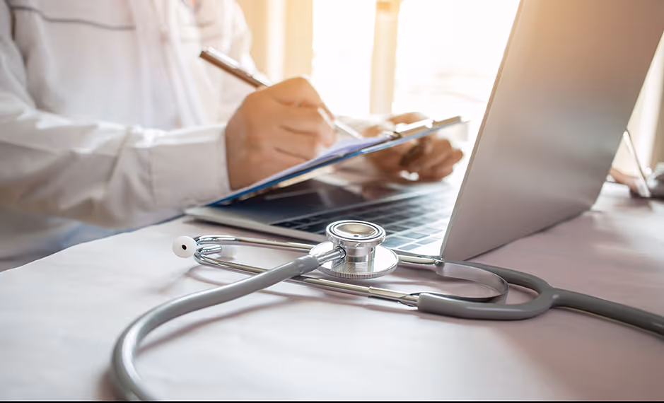 Doctor writes on clipboard beside laptop with stethoscope on table in bright office.