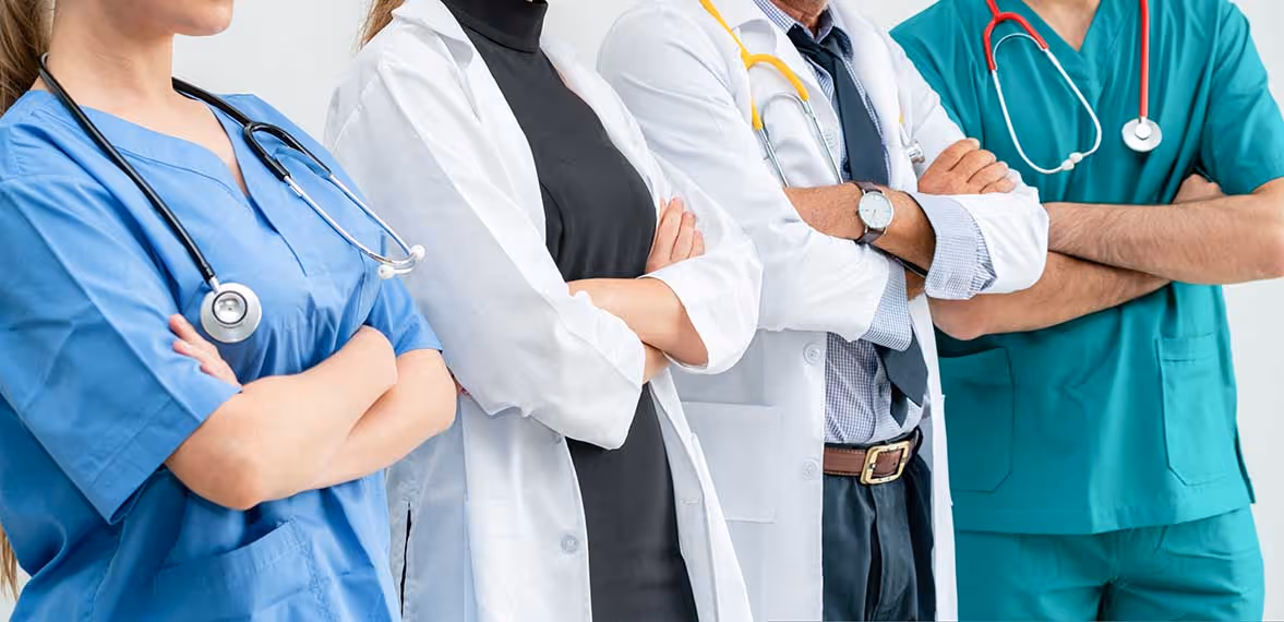 Four medical professionals with arms crossed wearing scrubs and white coats with stethoscopes.