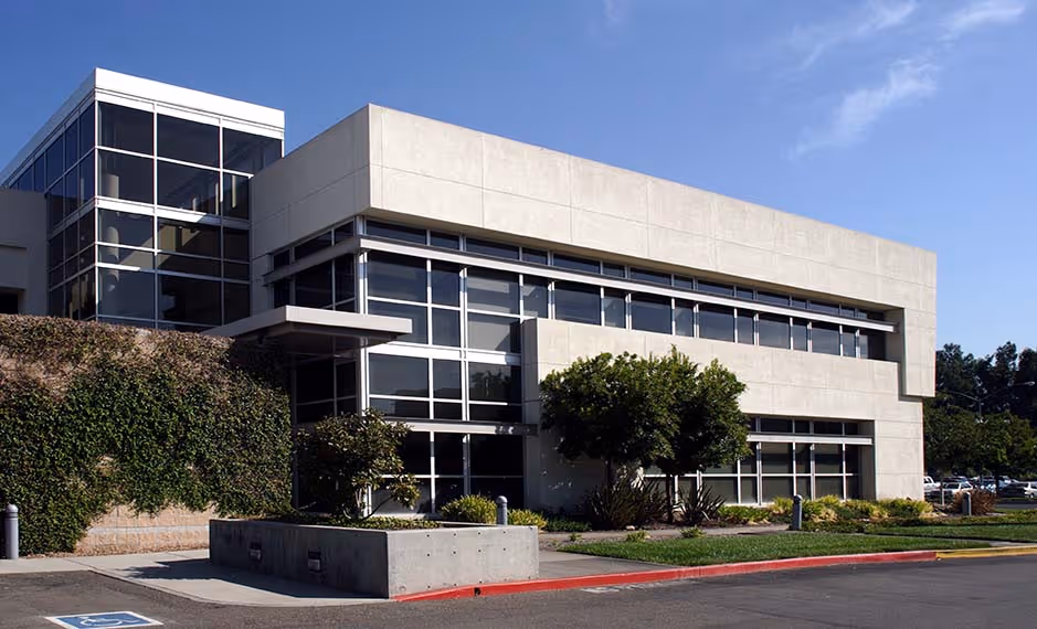 Modern two-story office building with large glass windows and surrounding greenery under a clear sky.