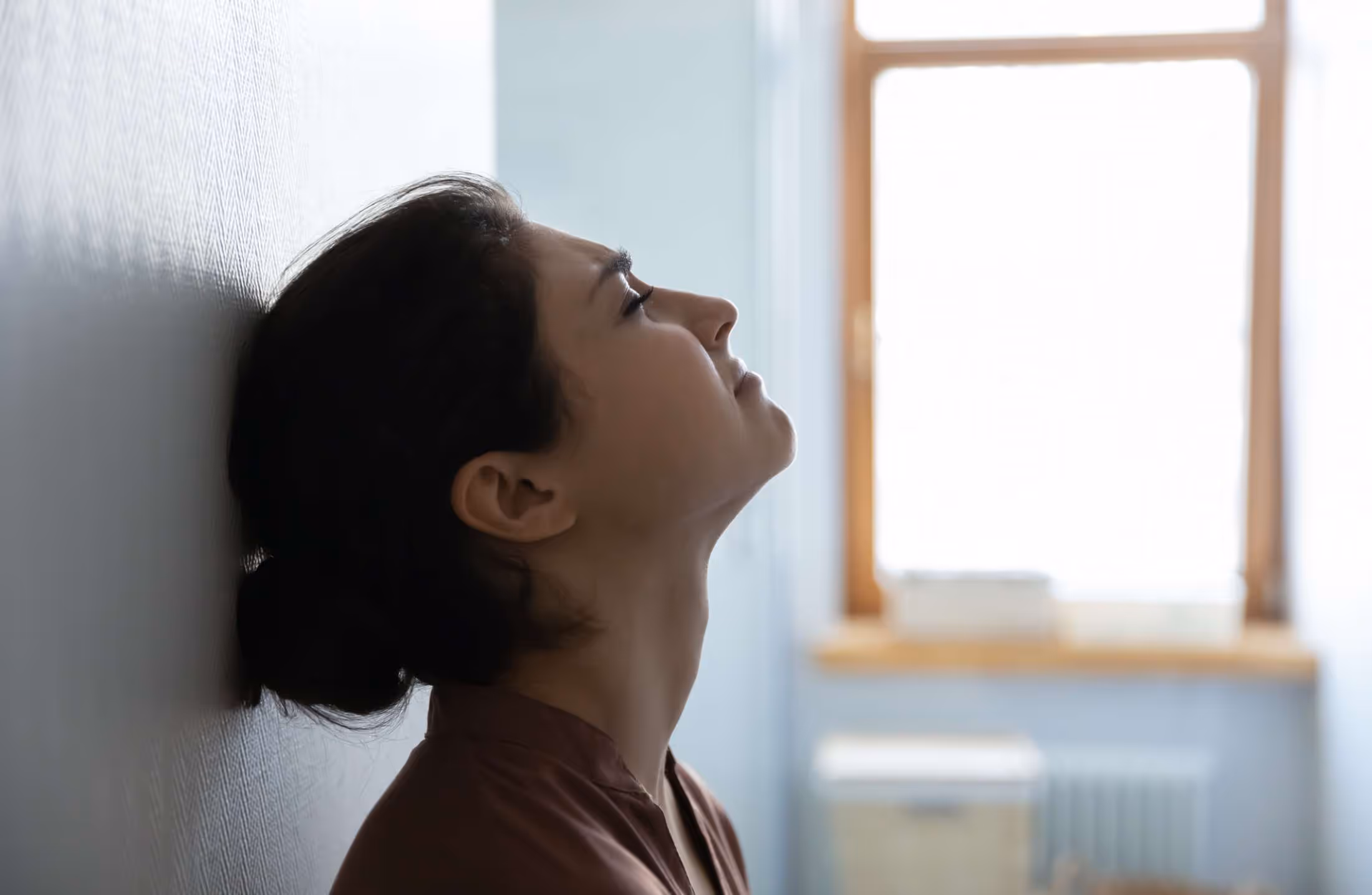 Side profile of a woman leaning her head against a wall looking upward, appearing thoughtful or stressed.