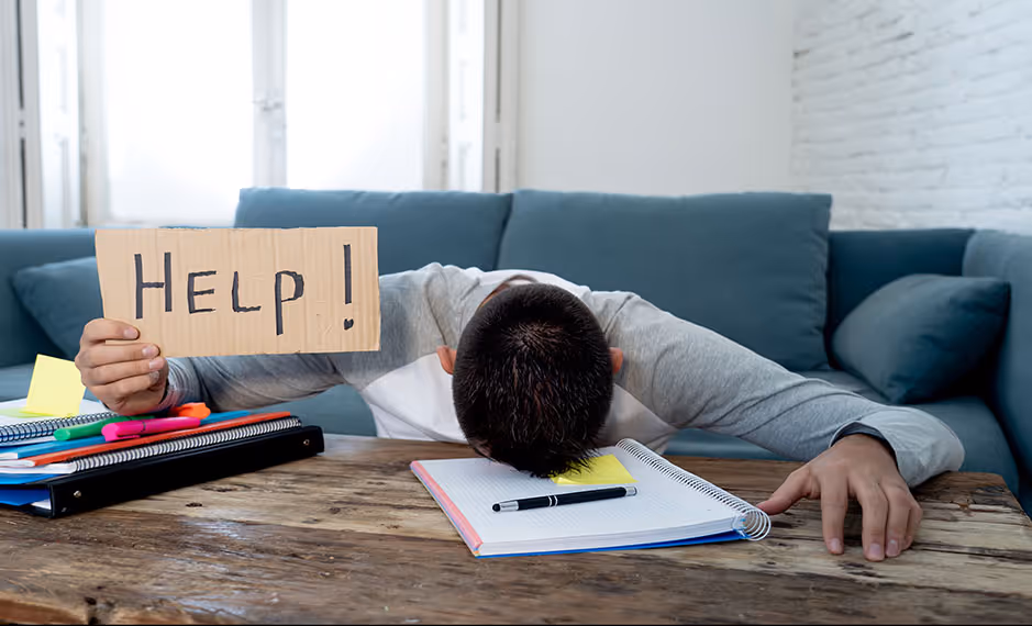 Frustrated person with head down on table holding cardboard sign reading 'HELP!' surrounded by notebooks and pens.