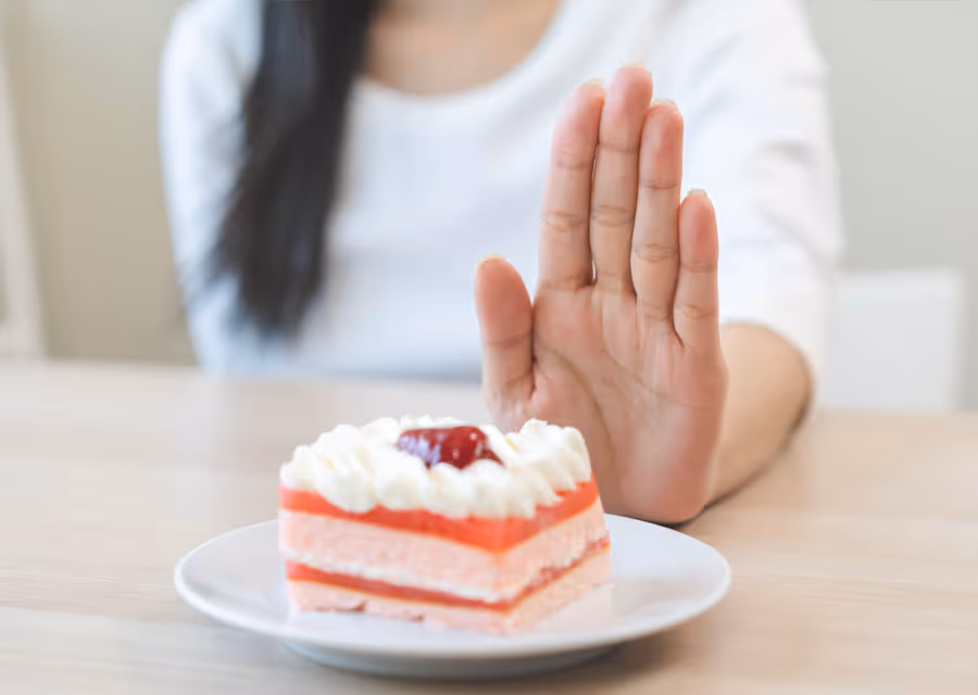 Person with raised hand in a stop gesture in front of a plate with a small pink and white layered cake.