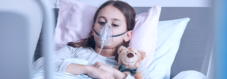 Young girl lying in bed with oxygen mask holding a teddy bear against pillows.