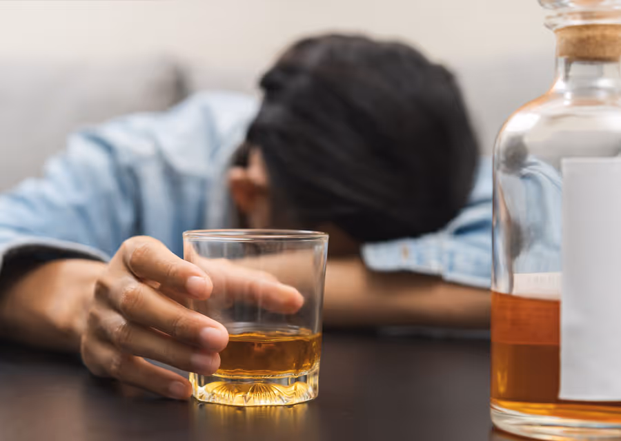 Person resting head on a table holding a glass of whiskey with a whiskey bottle nearby.