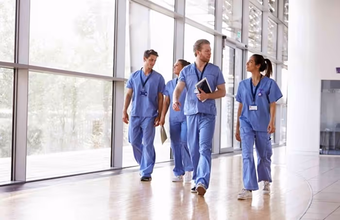 Four healthcare professionals in blue scrubs walking and talking in a bright hospital corridor.