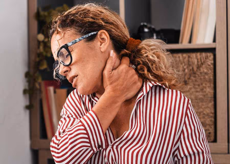Woman in striped shirt and glasses holding her neck with a pained expression indoors.