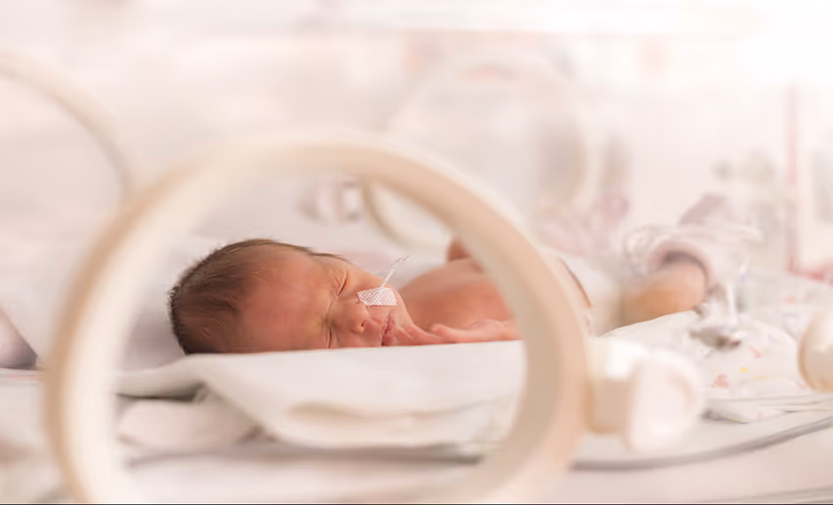 Premature newborn sleeping in an incubator with a feeding tube on the cheek.