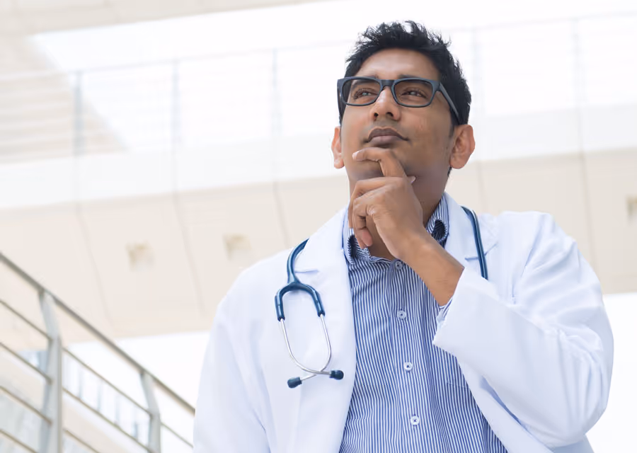 Thoughtful male doctor wearing glasses, white coat, and stethoscope in a bright modern building.