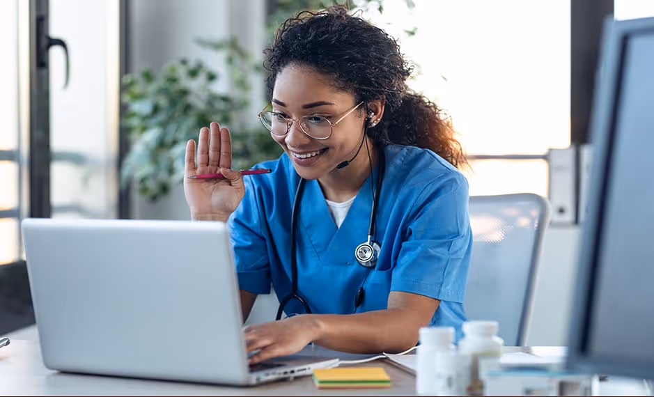 Smiling female nurse in blue scrubs with headset waving while using a laptop at her desk.