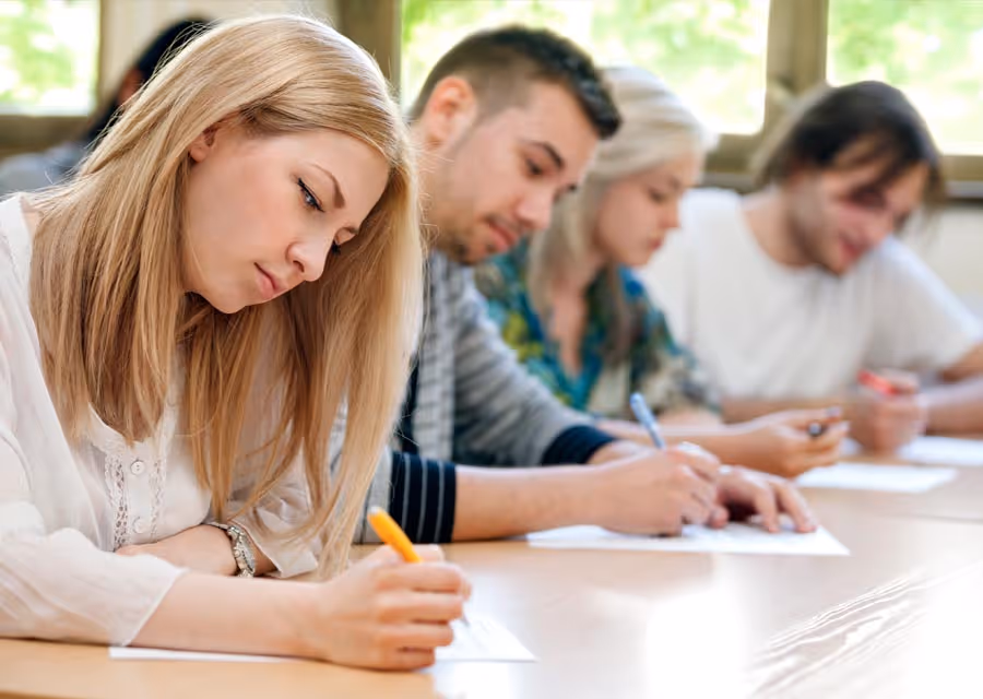 Four young adults sitting side by side writing on papers at a desk in a well-lit room.