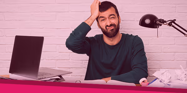 Man with beard scratching his head and wincing, sitting at desk with laptop and crumpled papers.