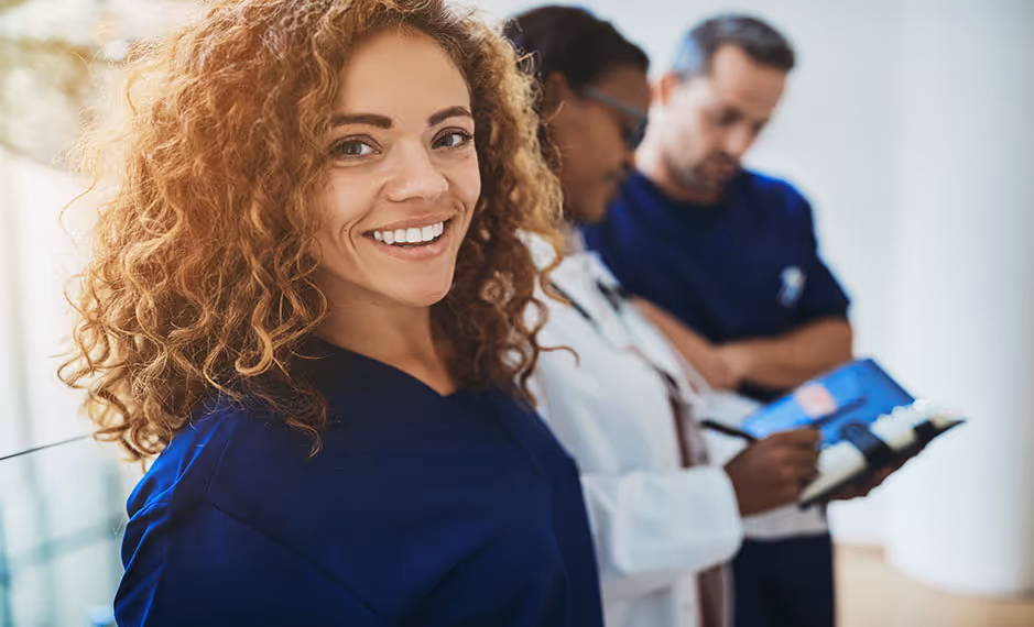 Smiling female healthcare worker in blue scrubs with two colleagues writing on clipboards behind her.