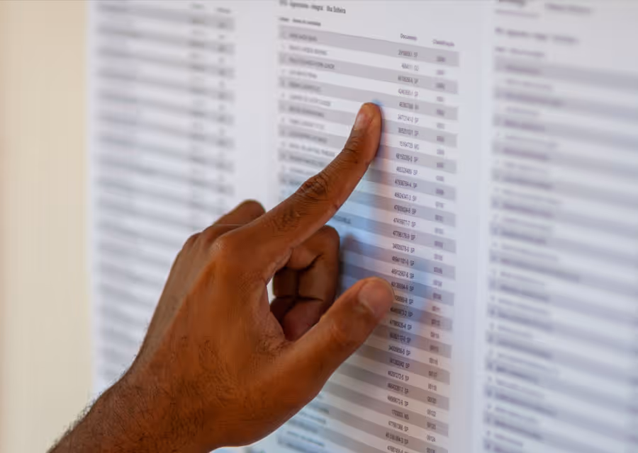 Hand pointing at an entry on a printed list of documents or data on white paper.