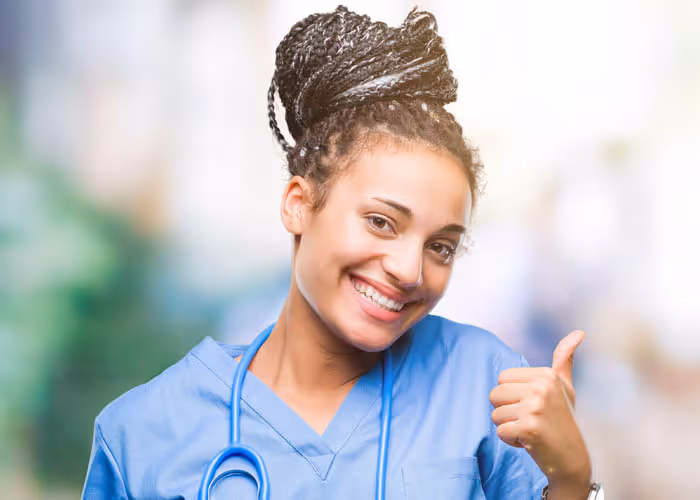 Smiling female healthcare worker in blue scrubs giving a thumbs-up with a stethoscope around her neck.