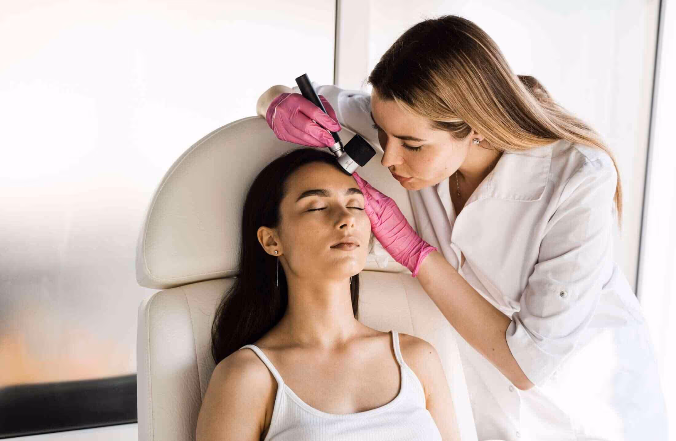 Dermatologist wearing pink gloves examines patient's forehead with dermatoscope in clinic.