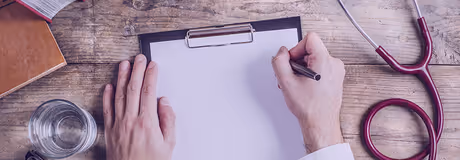 Person's hands on a clipboard with blank paper and a stethoscope on a wooden table.