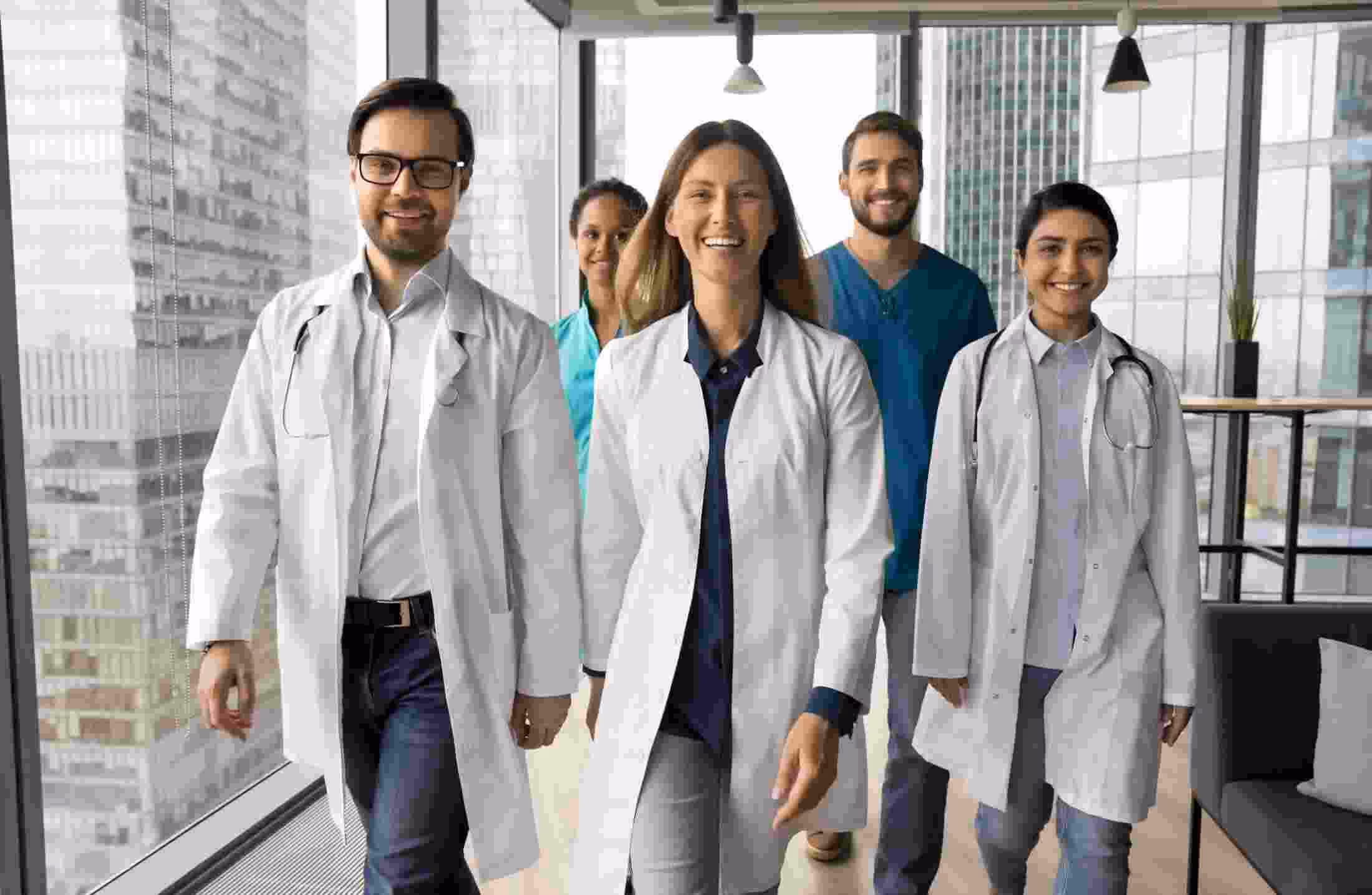 Group of five happy doctors and nurses walking confidently in a modern hospital hallway.