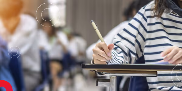 Person wearing striped shirt writing with pen on desk in a classroom setting with blurred students.