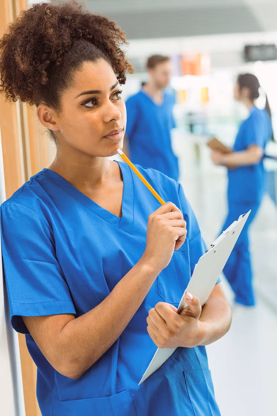 Thoughtful nurse in blue scrubs holds clipboard and pencil, with two colleagues blurred in background.