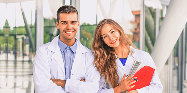 Smiling male and female medical professionals in white coats, standing indoors with clipboard and pen.