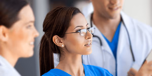 Three medical professionals in discussion, with one woman in blue scrubs and glasses in focus.