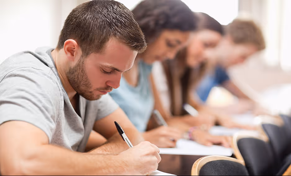 Four students seated at desks writing on paper in a classroom setting, focused on their work.