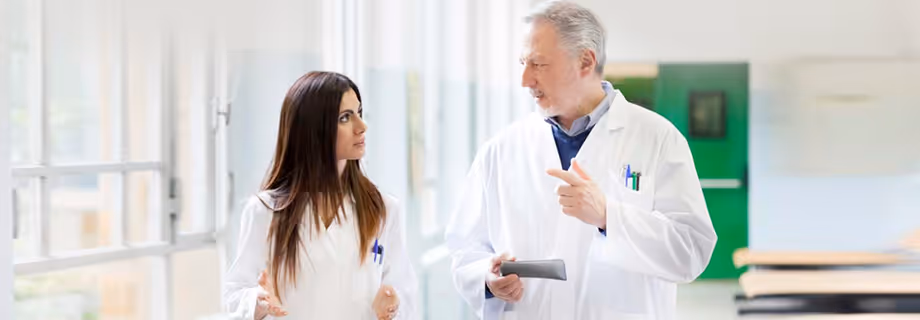 Two doctors in white coats having a discussion in a bright hospital corridor.