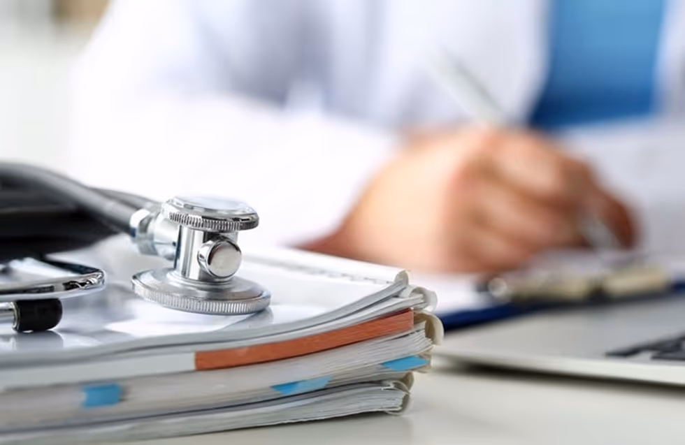 Stethoscope on stacked medical documents with a blurred person writing on clipboard in the background.