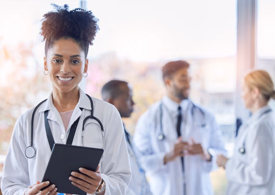 Smiling female doctor with curly hair holding a tablet, with three doctors conversing in background.