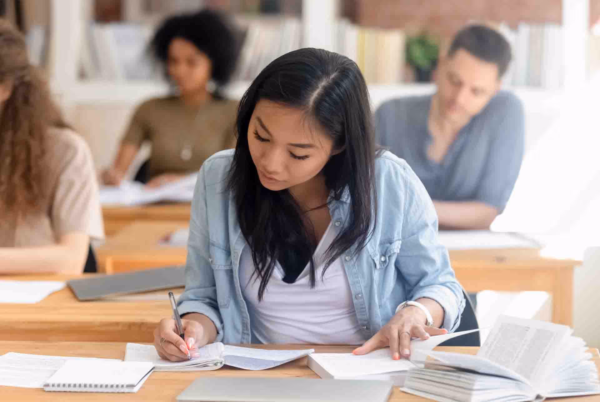 Female student writing notes while reading a book in a classroom with other students.