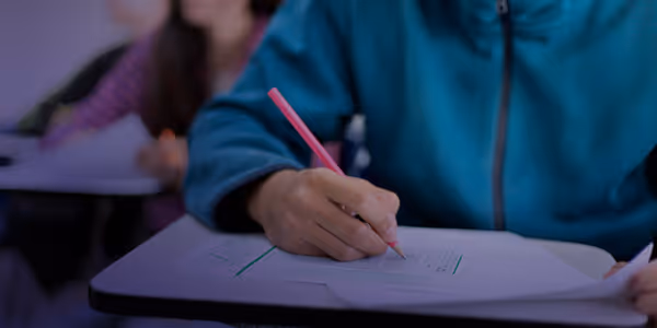 Person in blue jacket filling answer sheet with a pencil during an exam in a classroom.
