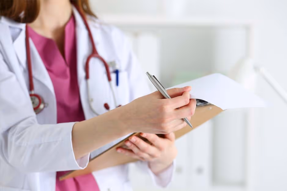 Medical professional in white coat with a stethoscope writing on a clipboard with a pen.